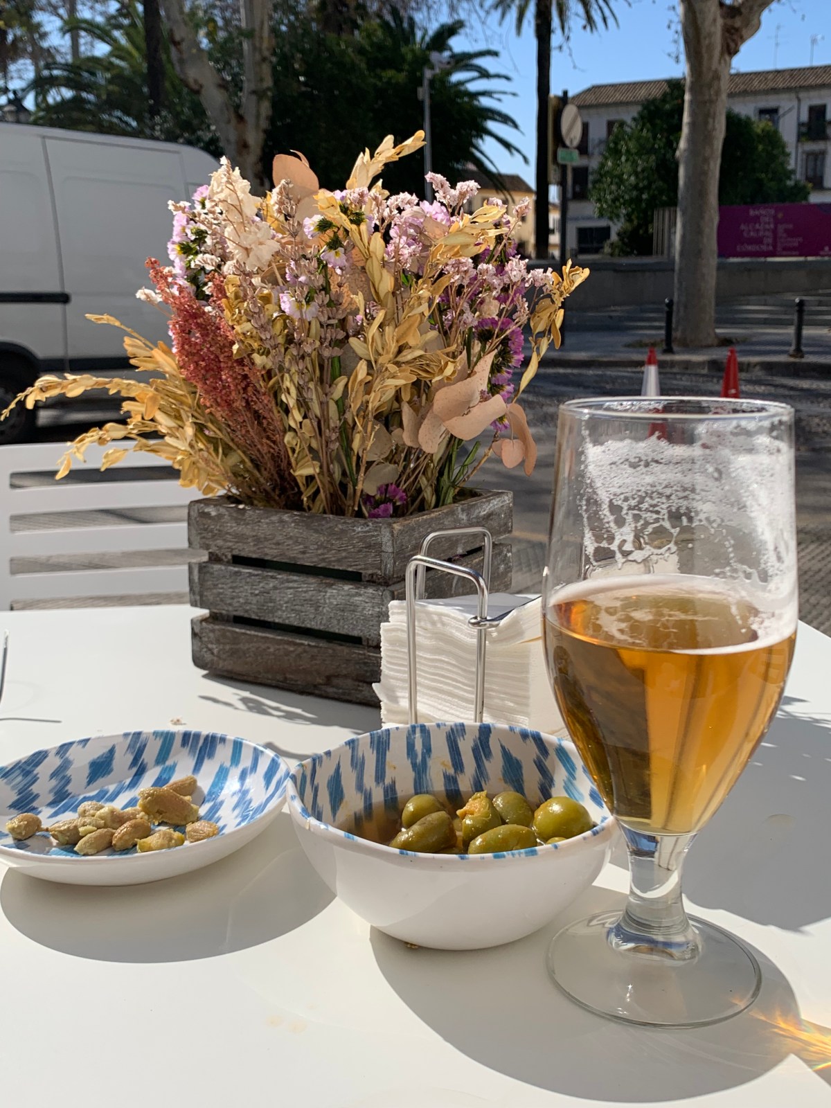 A table in the sunshine with green olives, olive pits, a glass of beer.
