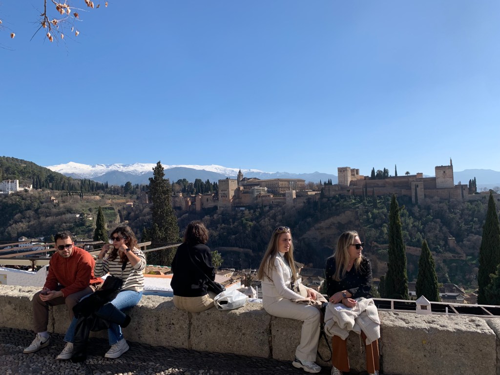 A vista overlooking the Alhambra and the Sierra Nevada mountains, with people sitting on a wall in the foreground.