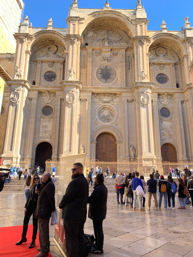 Front of large Cathedral with someone being interviewed on a red carpet and a security man in the foreground.