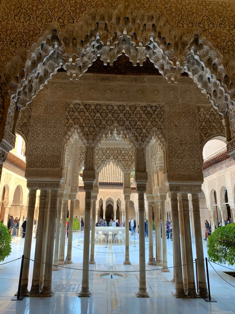 Columns supporting delicately carved arches, looking out to a plaza with a fountain surrounded by stone lions.
