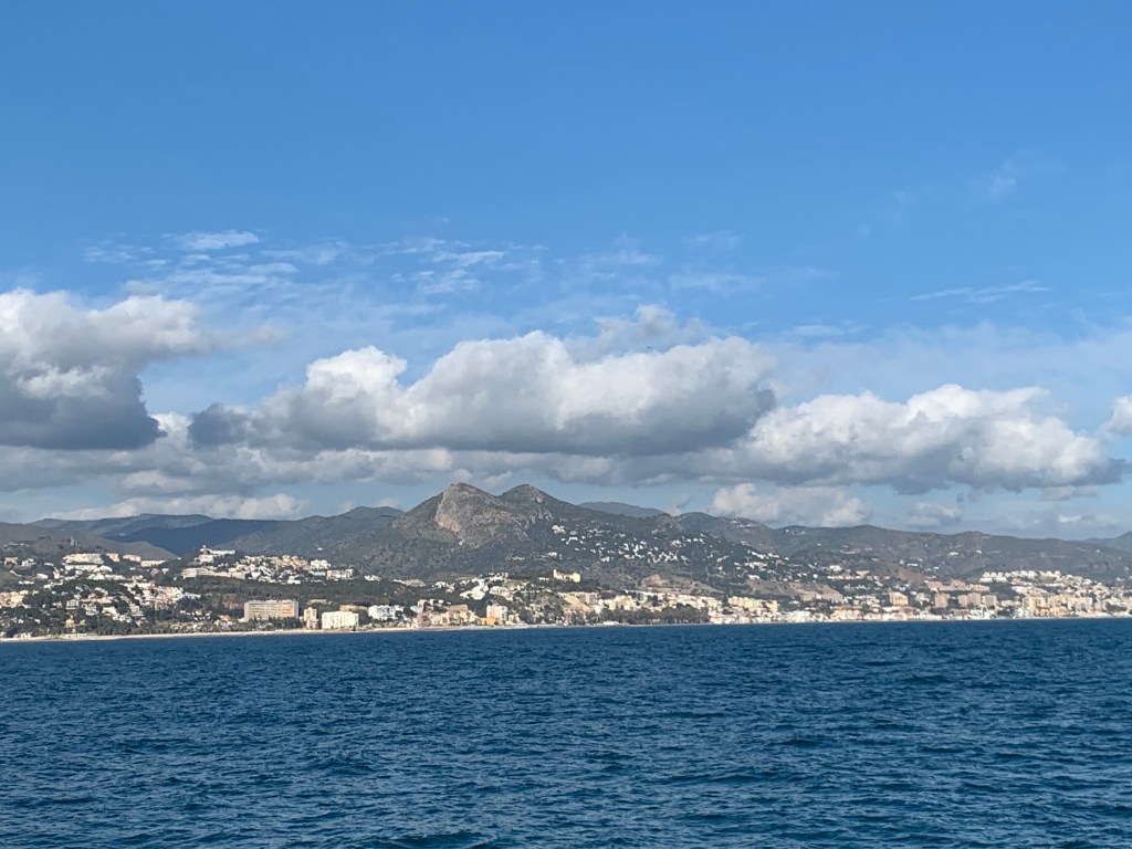 Coastline with white houses in the distance, a mountain range, and clouds dotting a blue sky.
