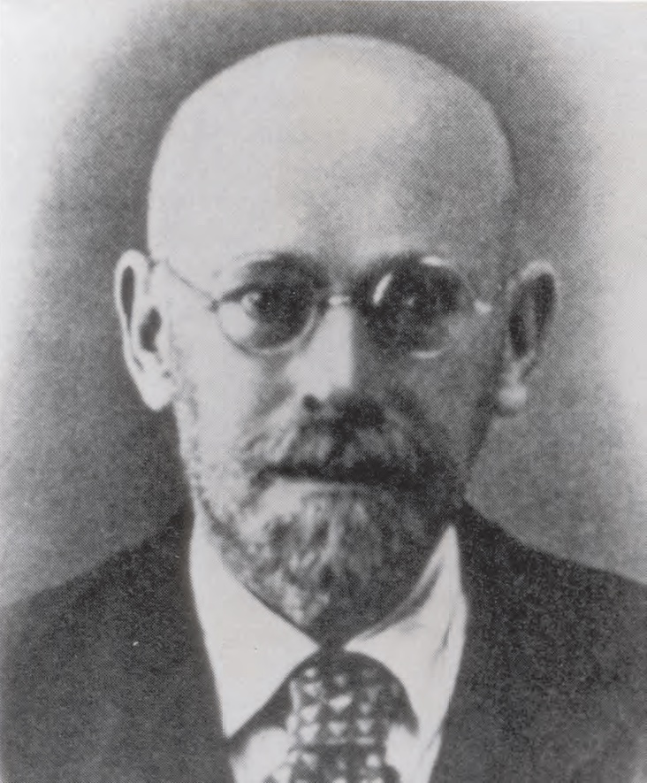 Photograph of Janusz Korczak, a bald man with a trim beard and moustache, wearing round glasses and a suit and tie.